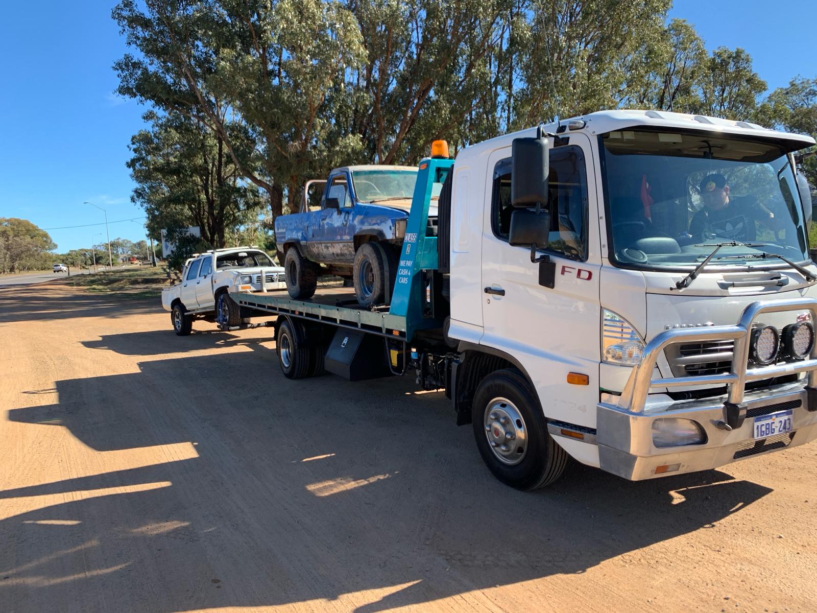 A white tow truck, pulling a silver car on a flatbed trailer with a yellow light on top, parked in front of houses with tr...